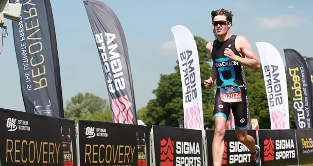 Male triathlete running past branded banners during a race, with Optimum Nutrition and Sigma Sports logos visible along the course.