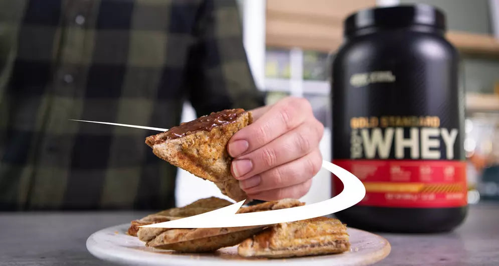 Proteinfylld jordnötssmör- och banantoast på en tallrik, en hand lyfter en bit, med ett kärl med vassleprotein i bakgrunden.