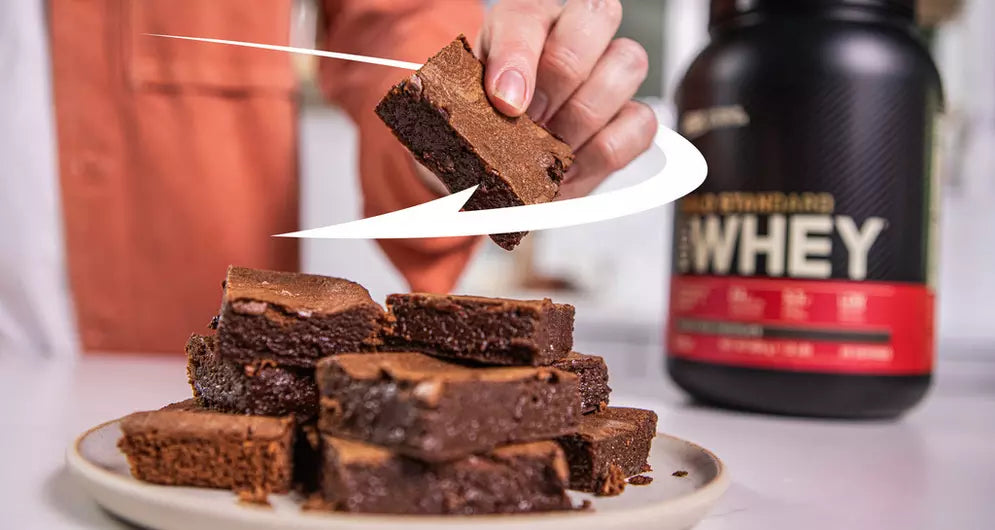 Chocolate protein brownies stacked on a plate, with a person lifting one brownie and a tub of whey protein in the background.