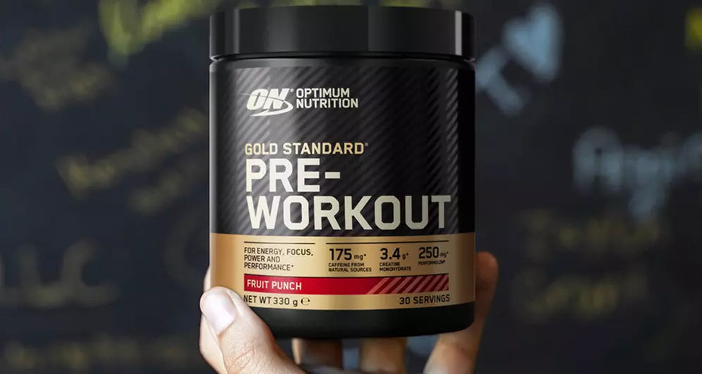 Hand holding a tub of Optimum Nutrition Gold Standard Pre-Workout Fruit Punch against a blurred dark background.