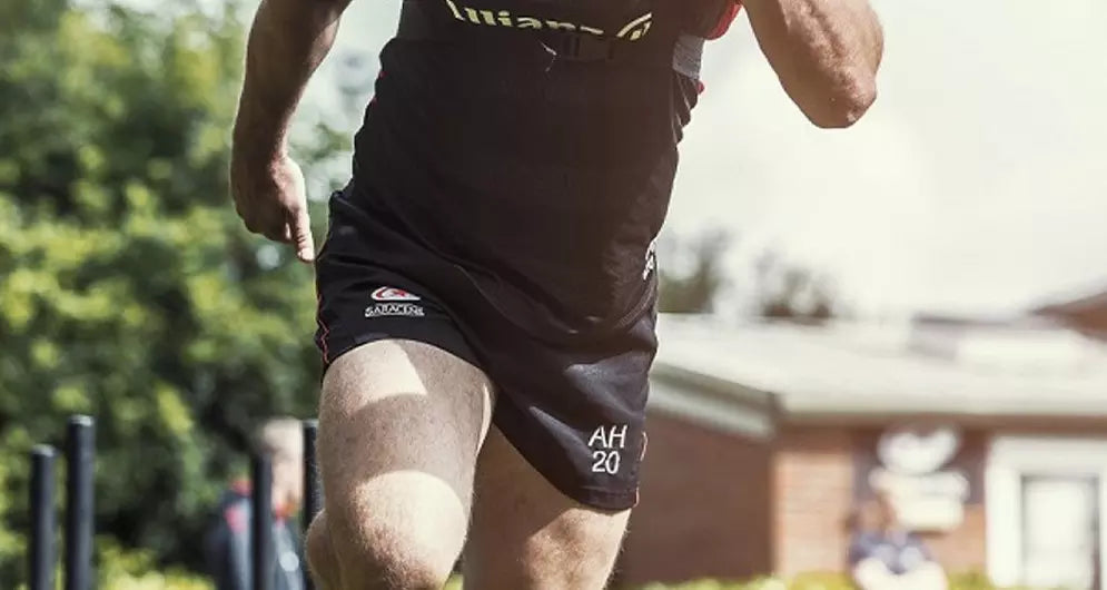 Close-up of a male athlete sprinting outdoors, wearing black training shorts with initials and a logo visible.