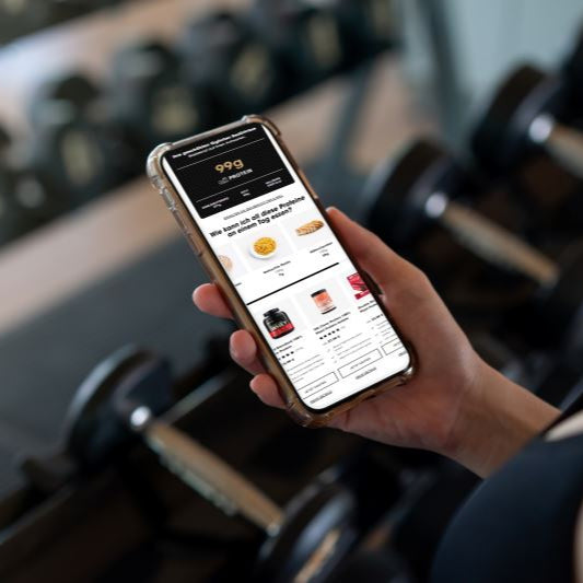 Person holding a smartphone displaying a fitness app with protein and supplement options, in a gym setting with dumbbells in the background