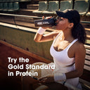 A woman in a white tennis outfit sits on stadium seating holding a protein shaker bottle with the Optimum Nutrition logo. Large text on the image reads 'Try the Gold Standard in Protein'.