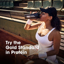 A woman in a white tennis outfit sits on stadium seating holding a protein shaker bottle with the Optimum Nutrition logo. Large text on the image reads 'Try the Gold Standard in Protein'.