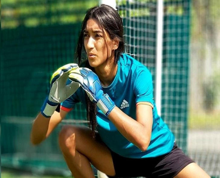 A person wearing a blue Adidas sports shirt and gloves, positioned in a goalkeeping stance on a soccer field