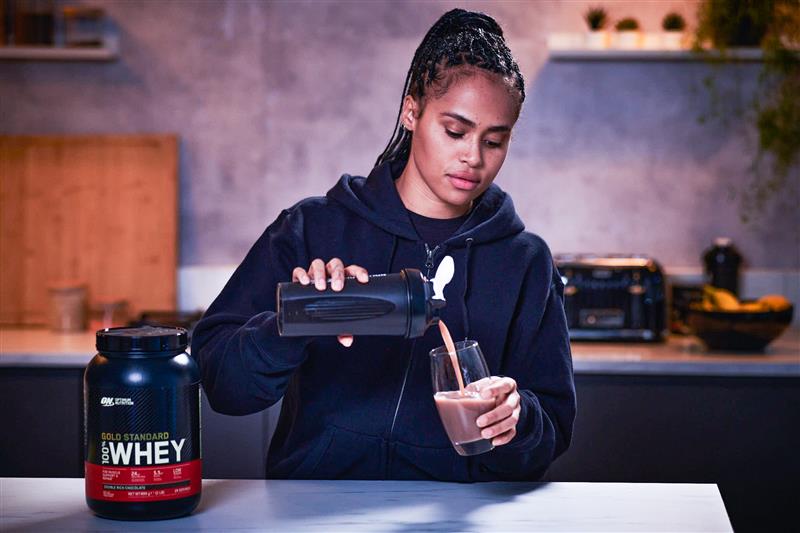 Optimum Nutrition Gold Standard 100% Whey Protein container next to a person pouring a protein shake into a glass