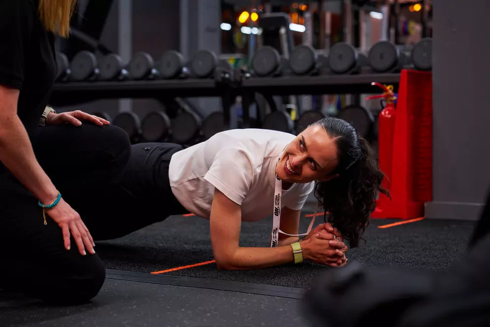A person is doing a plank exercise in a gym, assisted by a trainer. Dumbbells are visible in the background