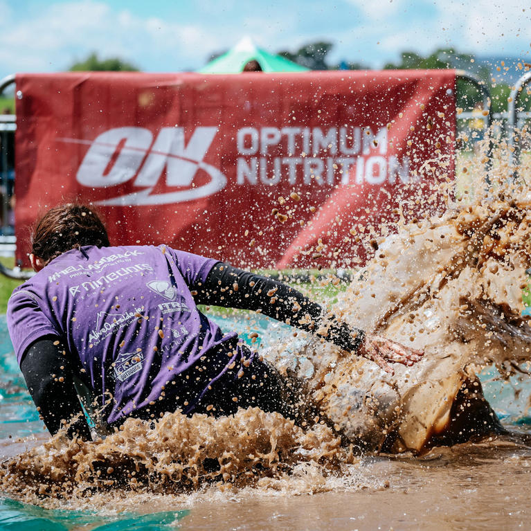 A person splashing through mud in front of an Optimum Nutrition promotional banner at an outdoor event