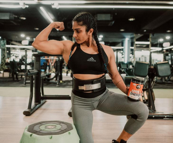 Person in a gym holding a container of Amino Energy formula, wearing workout attire, flexing arm
