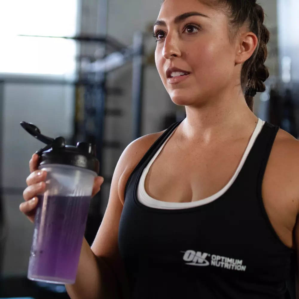 Optimum Nutrition shaker bottle held by a person in a gym setting, wearing an Optimum Nutrition tank top