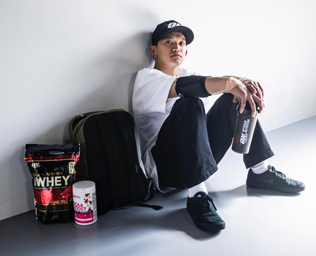 Optimum Nutrition Gold Standard 100% Whey Protein, 2.73 kg bag, and BCAA Powder Raspberry Pomegranate flavour container next to a seated per