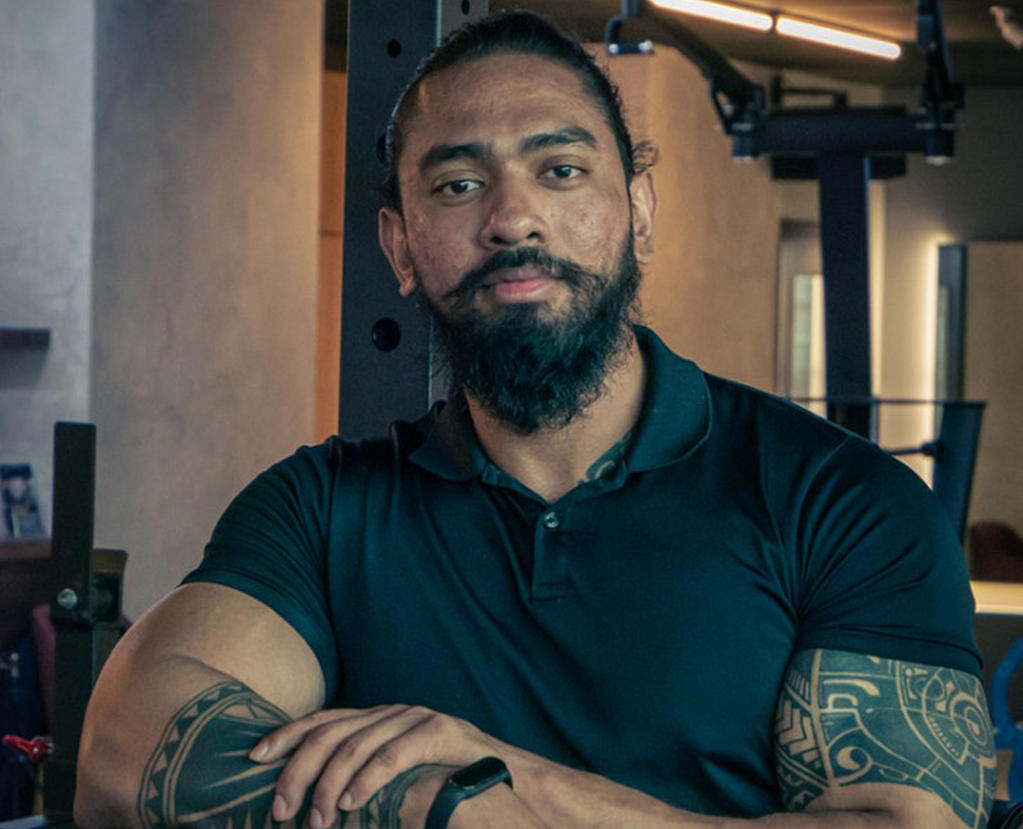 A person standing in a gym setting, wearing a black polo shirt, with gym equipment in the background