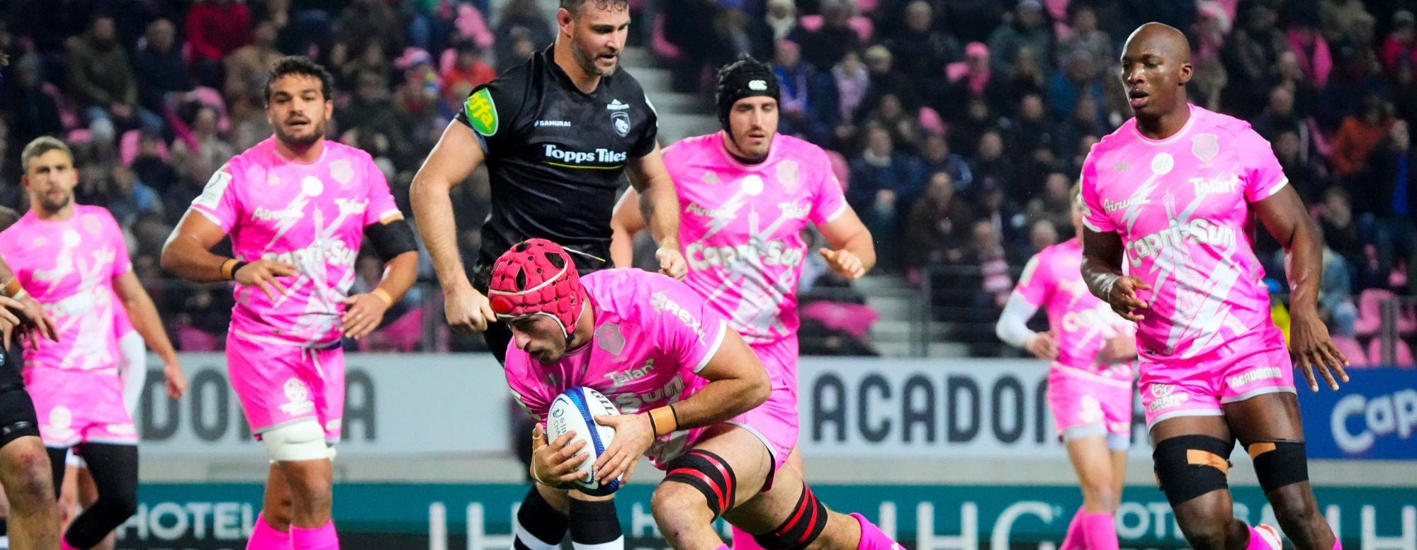 Rugby players in action during a match, with one player holding the ball while others prepare to engage