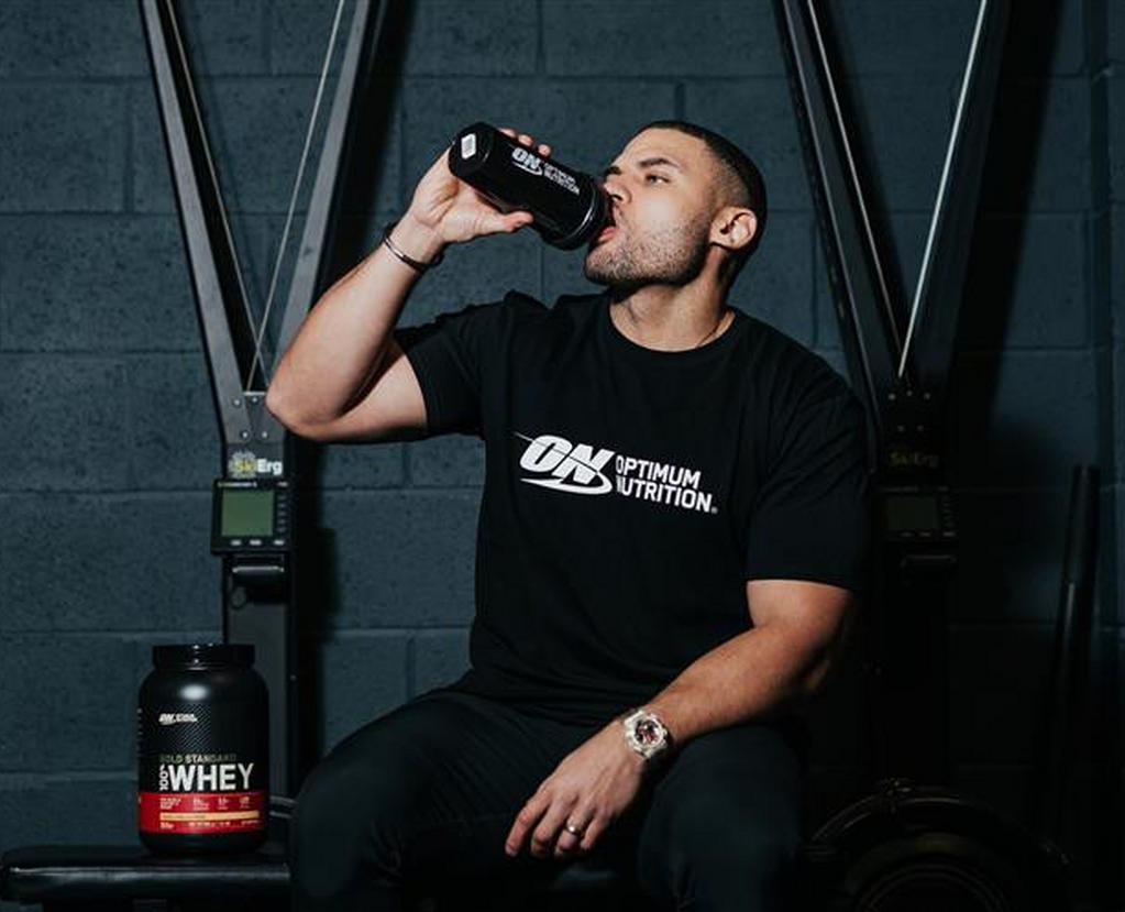 Man drinking from Optimum Nutrition shaker, seated next to Optimum Nutrition Gold Standard 100% Whey Protein container