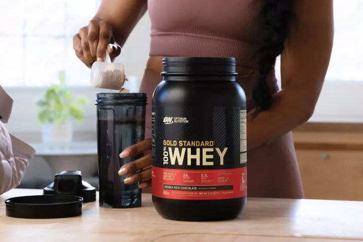 Optimum Nutrition Gold Standard 100% Whey Protein, 2.73 kg container, with person scooping powder into a shaker bottle