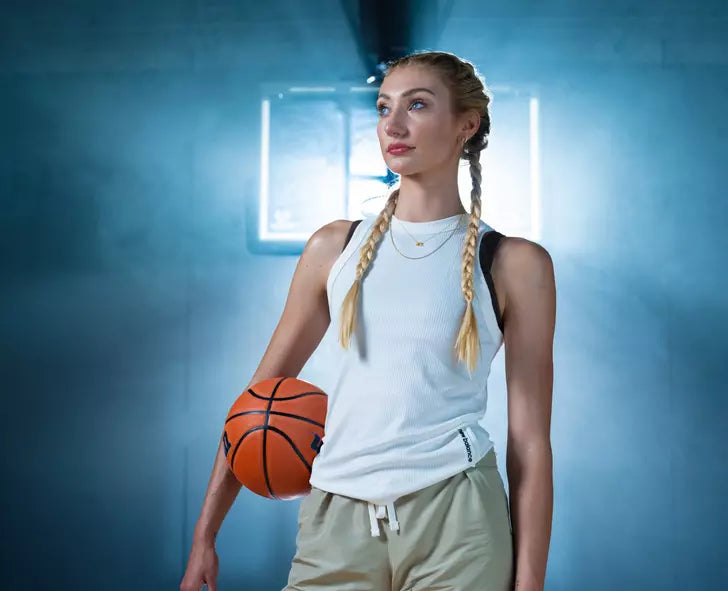 A woman holding a basketball, wearing a white tank top and beige pants, standing in a sports setting