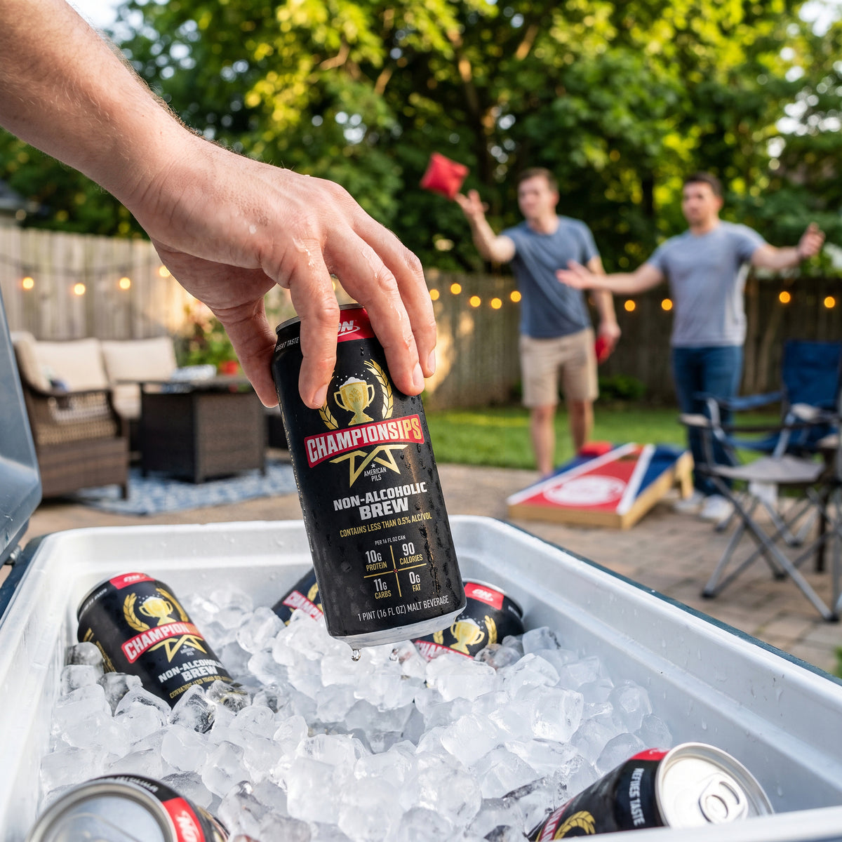 A hand holds a can of Champion's Non-Alcoholic Brew beer in front of an open cooler filled with ice, with two men playing backyard games in a sunny, wooded backyard setting.