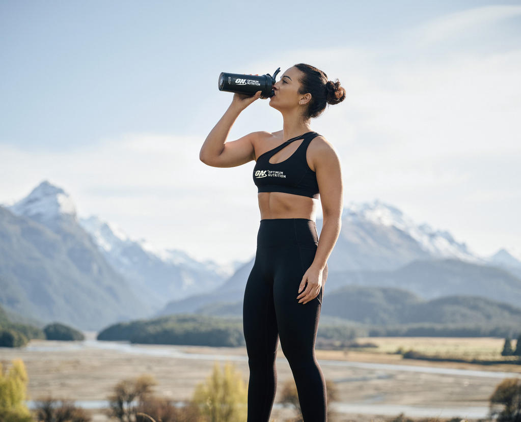 A woman drinks from a shaker bottle labeled "ON" in a mountainous outdoor setting