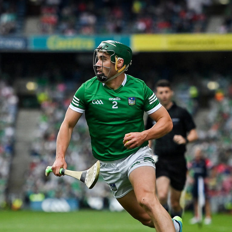 A hurler in action on the field, wearing a green jersey and helmet, holding a hurley stick