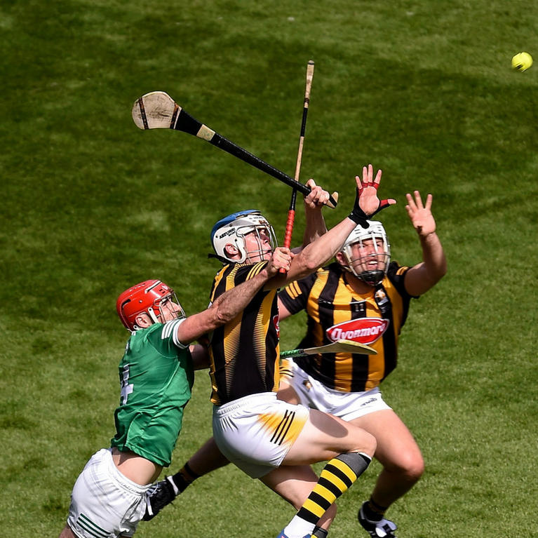 Three hurling players in action on a grass field, competing for the ball mid-air