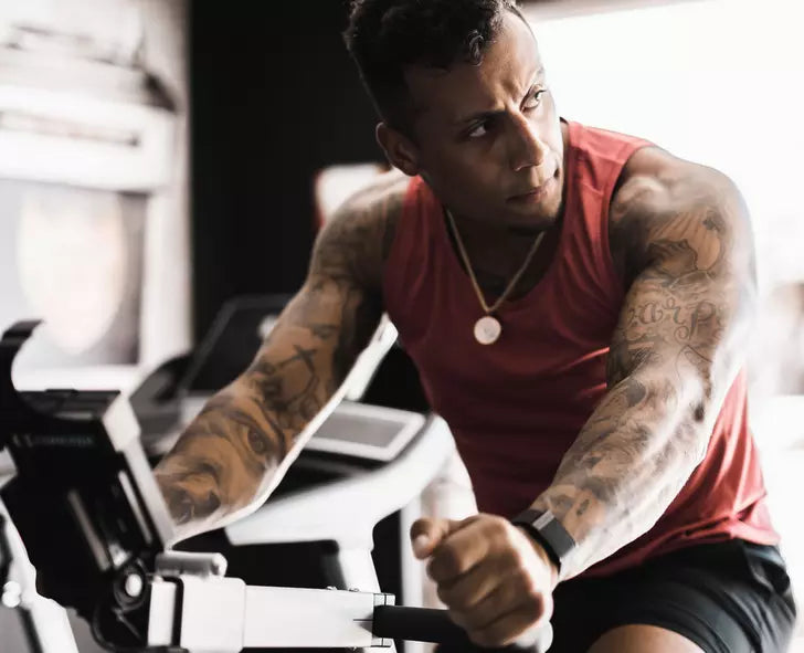 Man exercising on a stationary bike indoors, wearing a red tank top and fitness tracker
