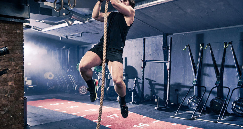Person climbing a rope in a gym setting, surrounded by exercise equipment