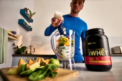 Man adding powder to blender with fruits and vegetables; Gold Standard 100% Whey protein container on counter
