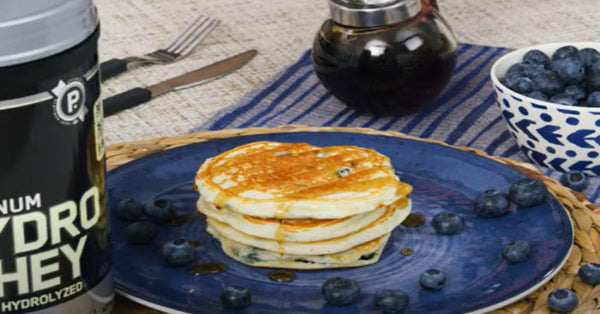 Stack of pancakes with syrup and blueberries on a plate, next to a container of protein powder