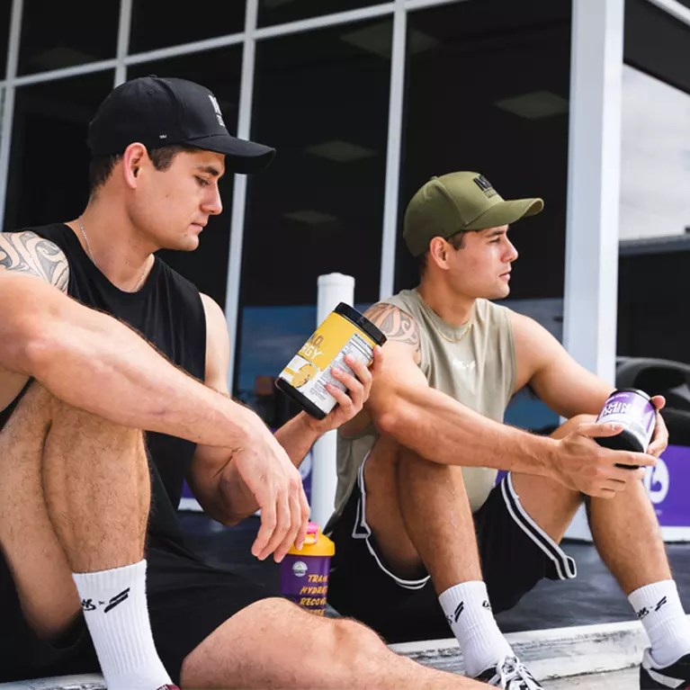 Two people sitting outside a building, each holding a container of nutritional supplements