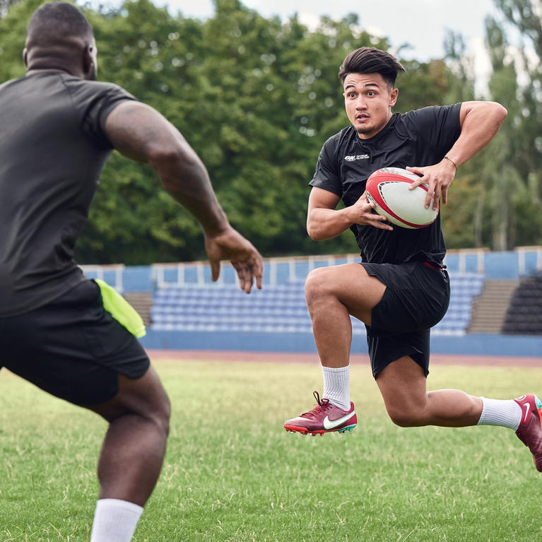 Two athletes playing rugby on a field, one running with the ball while the other prepares to tackle