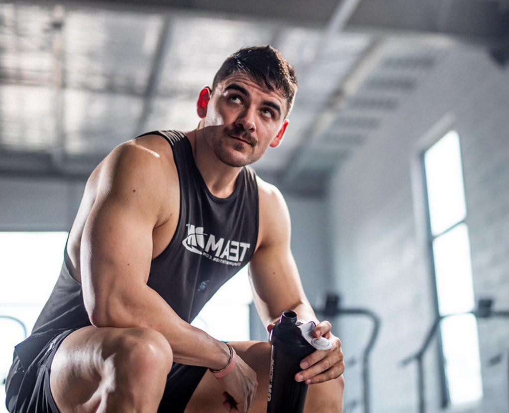 A man in a gym holding a protein shaker bottle, wearing a sleeveless shirt
