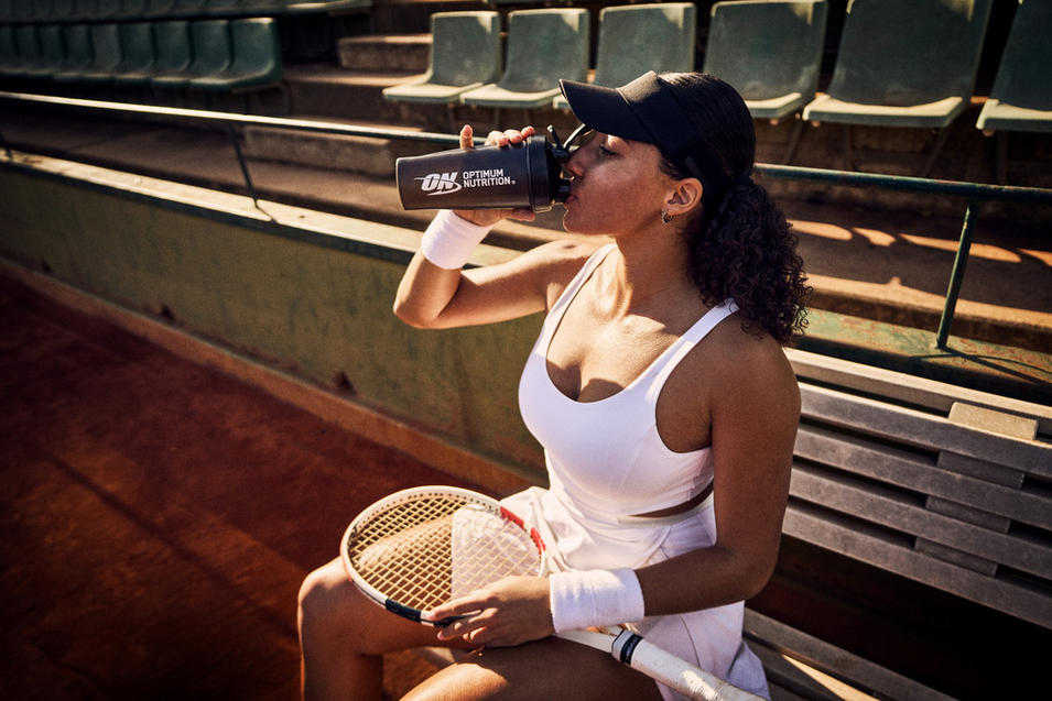 Person drinking from an Optimum Nutrition shaker bottle while sitting on a tennis court bench