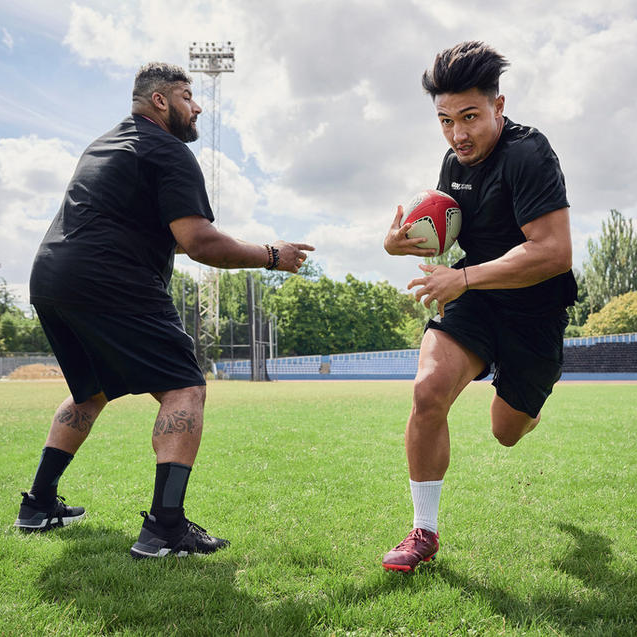 Two men playing rugby on a grassy field, one holding a rugby ball and running while the other attempts to tackle