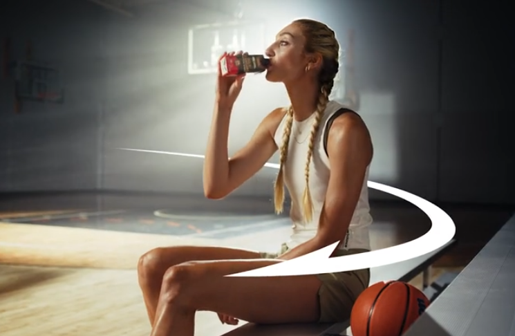Person drinking a protein shake in a gym setting, seated on a bench next to a basketball