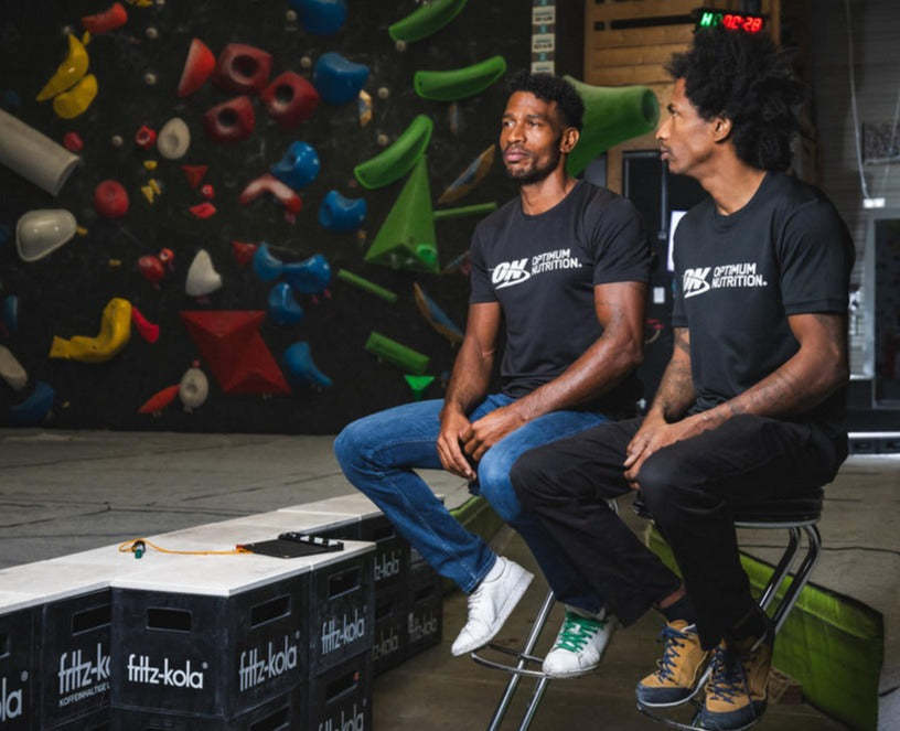 Two individuals wearing Optimum Nutrition branded shirts sitting on stools in an indoor climbing gym