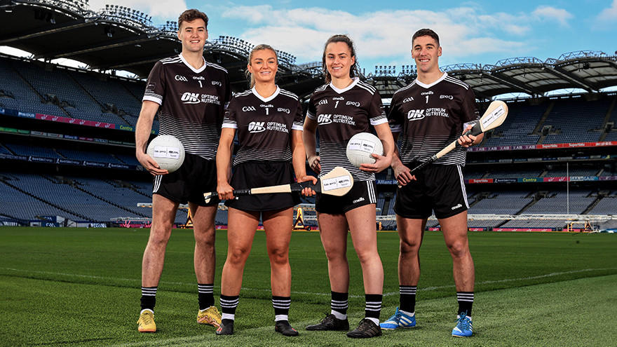 Four athletes in black sports uniforms stand on a sports field, holding Gaelic footballs and hurleys