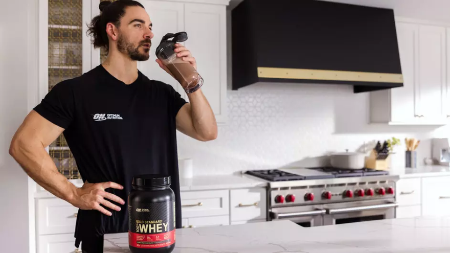 Man drinking a protein shake in a kitchen with a container of Gold Standard 100% Whey protein powder on the counter