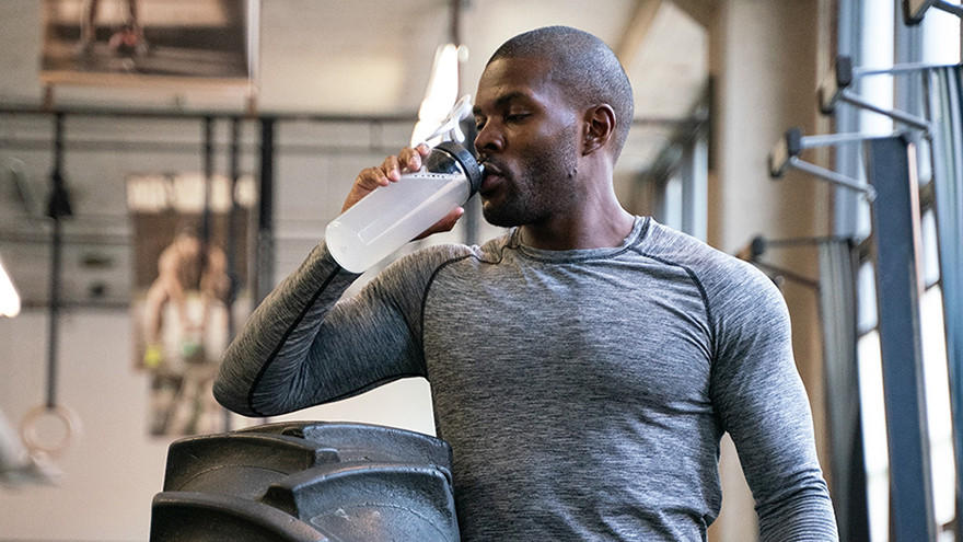 Man drinking from a shaker bottle in a gym setting, possibly post-workout