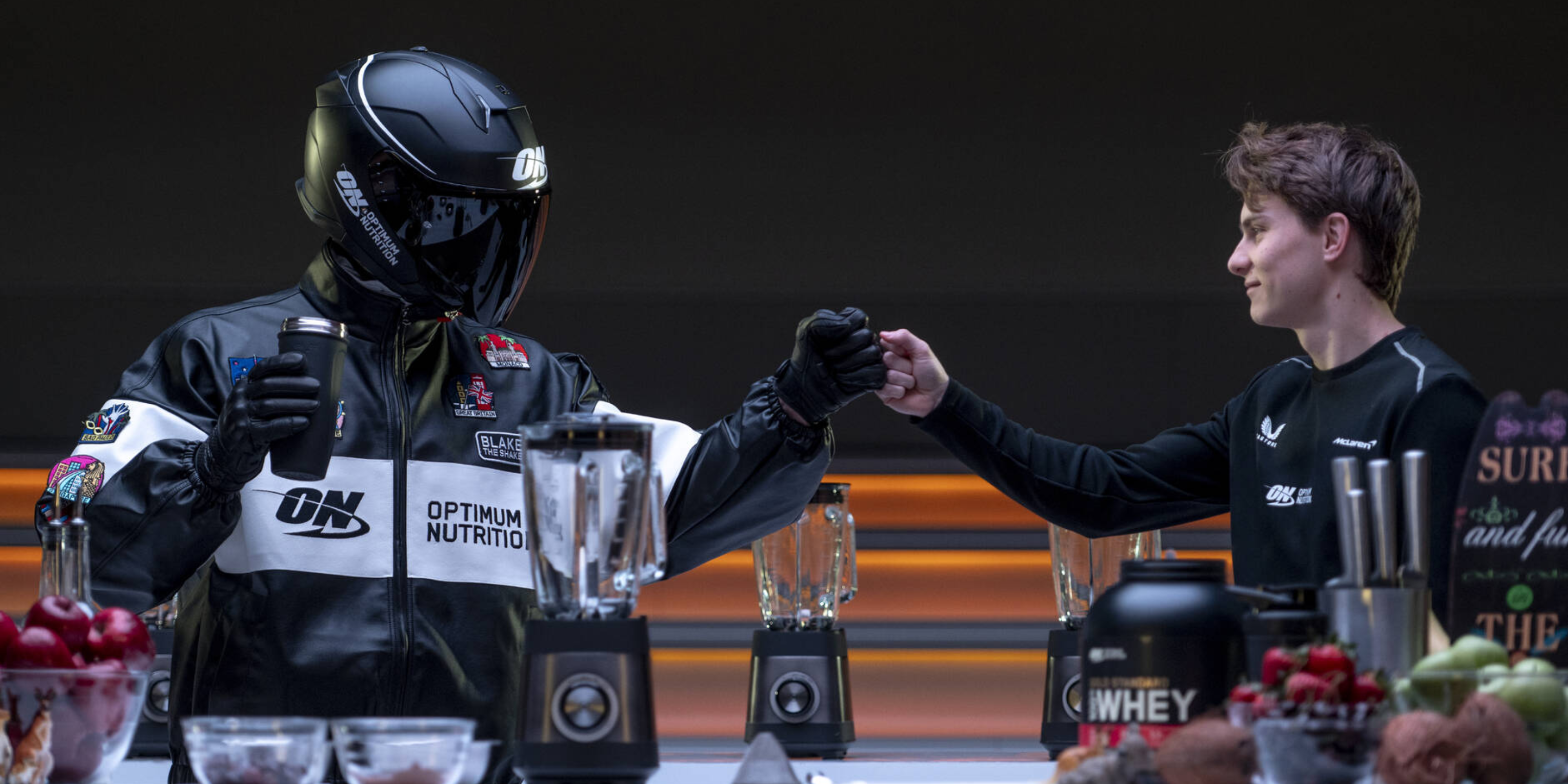 Two people in racing gear with blenders and Optimum Nutrition products on a table.