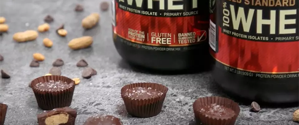 Optimum Nutrition Gold Standard 100% Whey Protein containers with chocolate pieces and nuts scattered around