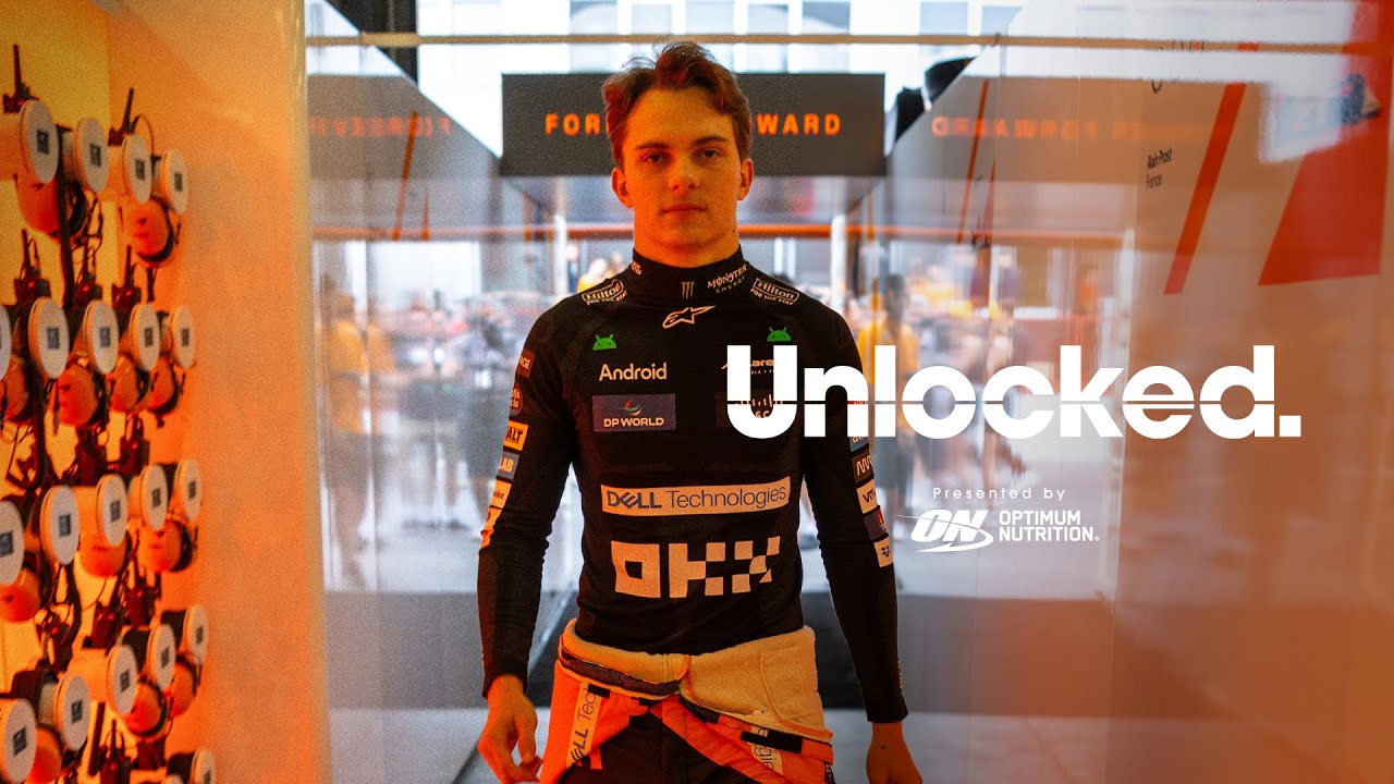 A person in racing gear stands in a technology-themed setting with "Unlocked" and "Optimum Nutrition" branding visible