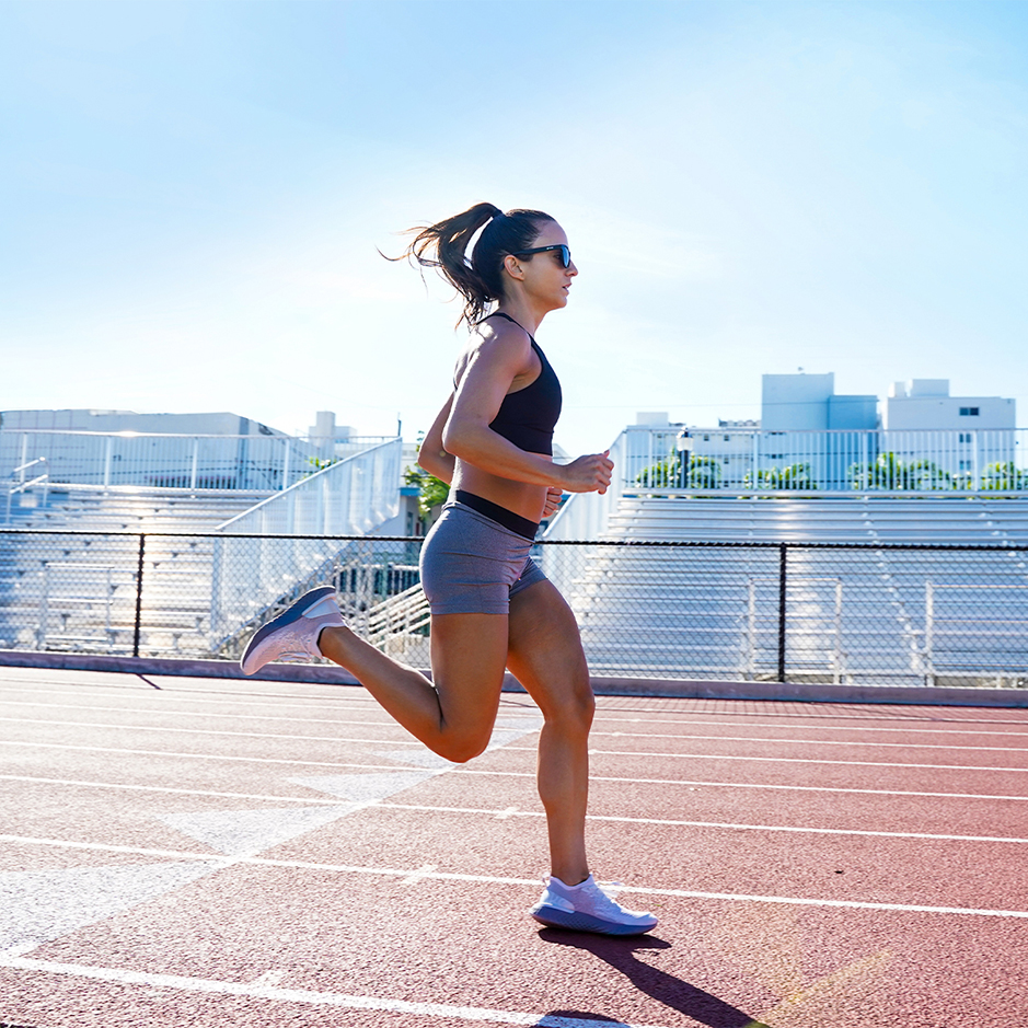 woman running on a track pitch outside in sunshine with sunglasses on
